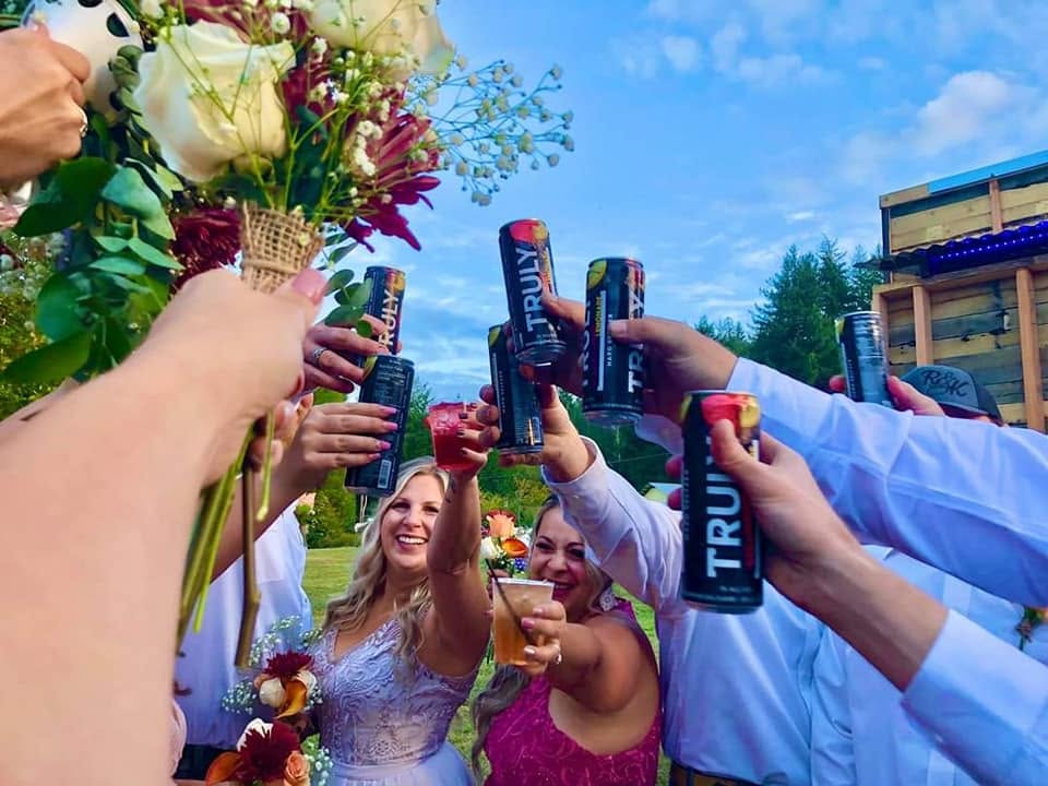 A wedding toast outdoors under a blue sky, the bride and her guests laughing as they raise drinks together in a circle, with a bridal bouquet held up in the foreground.
